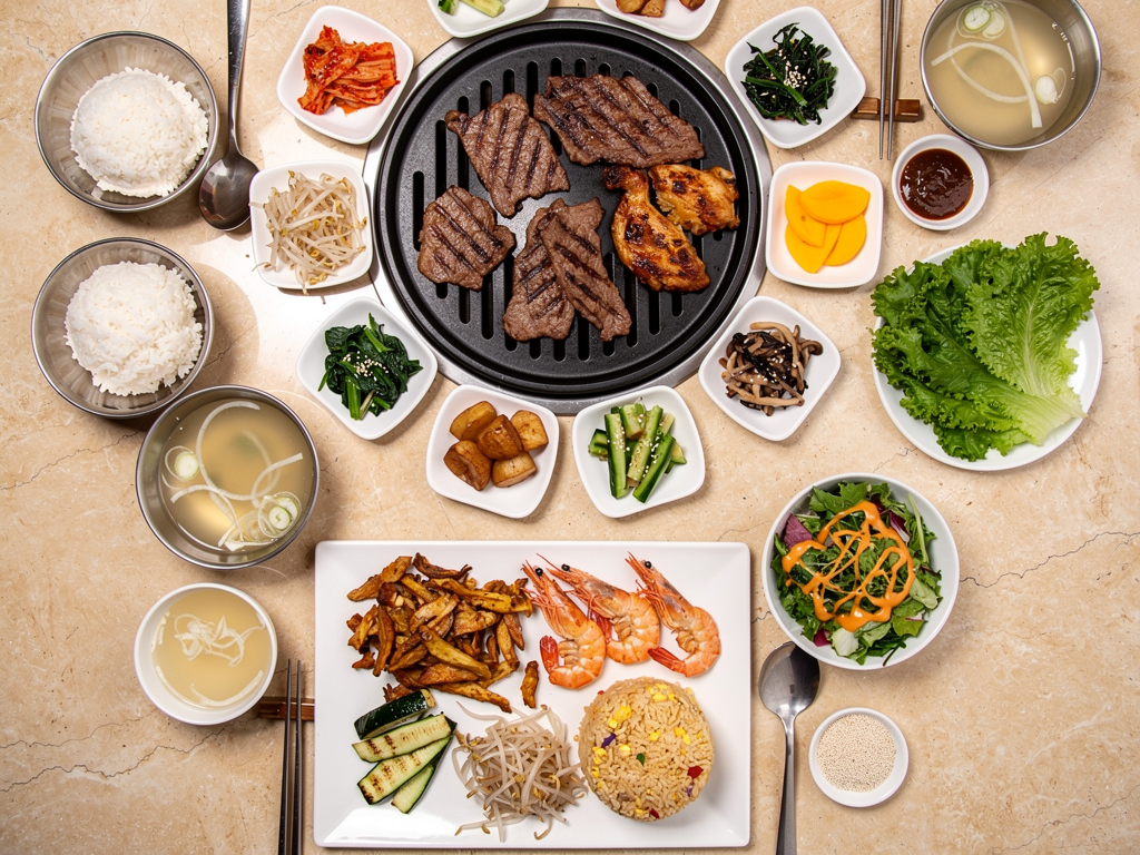 Mukbang Shows Korean BBQ table with beef, chicken, banchan, rice and soup beside a teppanyaki course on a white plate